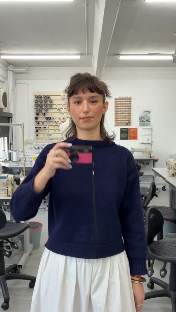 A person with short dark hair wearing a navy sweater and white skirt stands in a sewing workshop, holding up a pink and black camera, looking directly at the viewer. Sewing machines and tools are visible in the background.