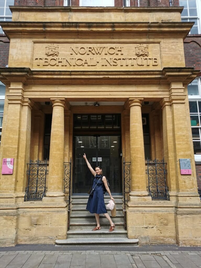 A person in a blue dress stands on the steps of Norwich Technical Institute, smiling and pointing upward at the building’s sign, with large columns and a decorative stone entrance behind them.