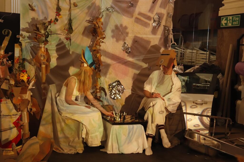 Two people in whimsical costumes, one wearing a blue crown and the other a cardboard box helmet, sit across from each other playing chess in an artfully decorated, surreal setting with cardboard and metallic decorations.