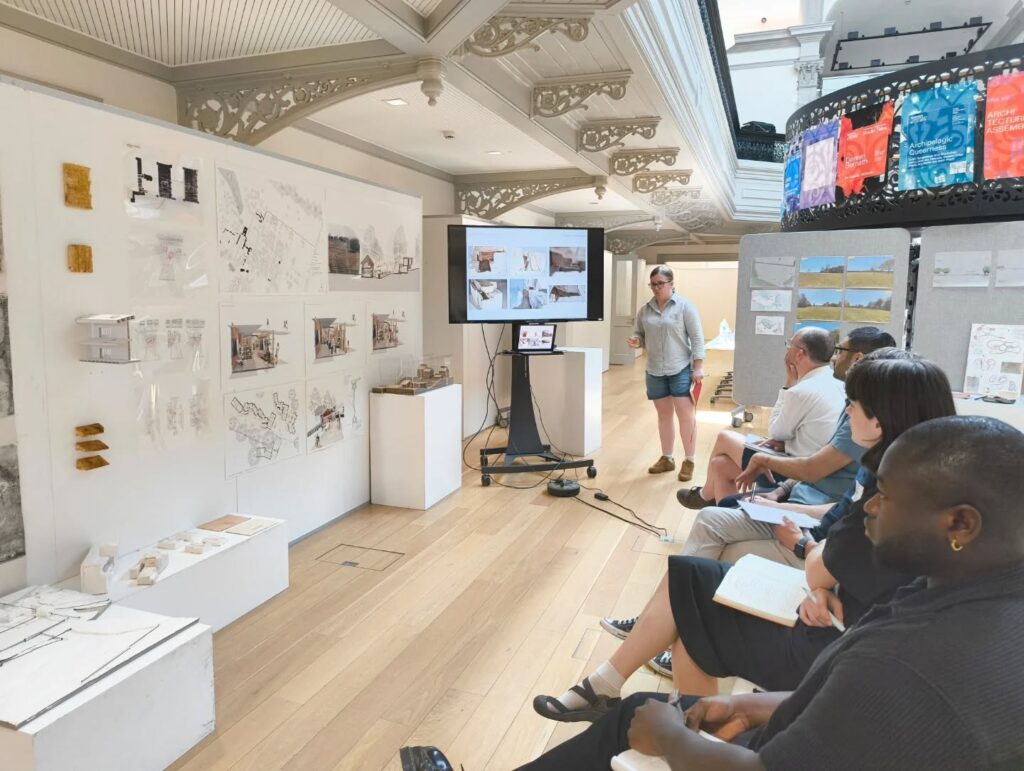A person gives a presentation in a bright, ornate room, standing by a screen showing architectural designs. Several people sit opposite, observing and taking notes. Design boards and models are displayed on the walls and tables.