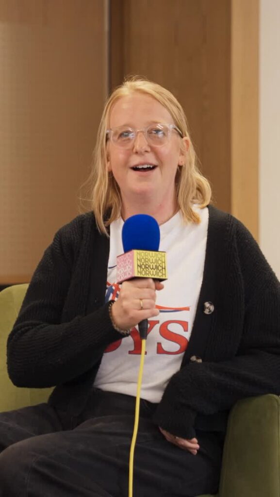 A person with long blonde hair and glasses smiles while holding a blue and yellow microphone, sitting on a green chair and wearing a black cardigan over a white T-shirt.