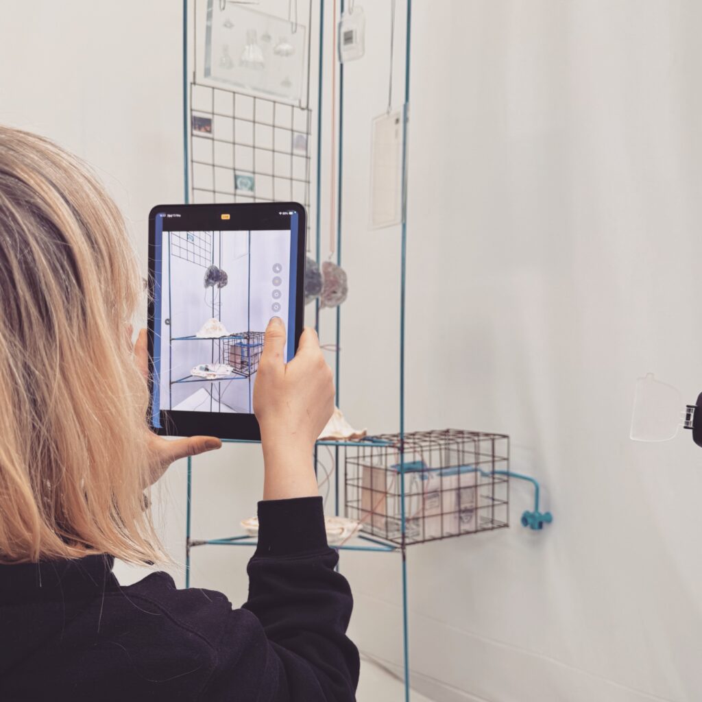 A person with blonde hair holds a tablet, taking a photo of a modern art display with wire shelves and small sculptures, inside a white-walled gallery space.