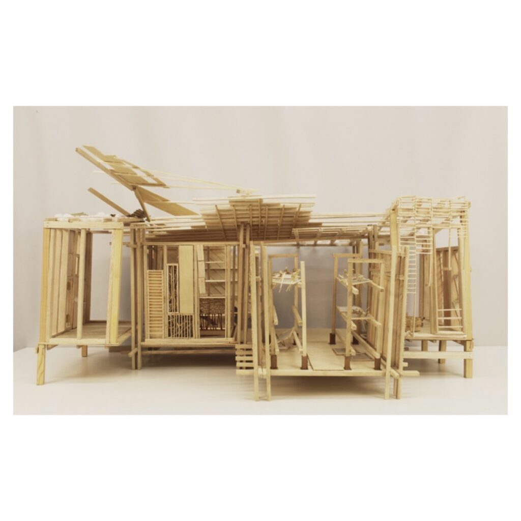 A detailed architectural model made of light wood, featuring open-framework walls, layered roof elements at varying angles, and multiple interconnected sections, displayed against a plain white background.