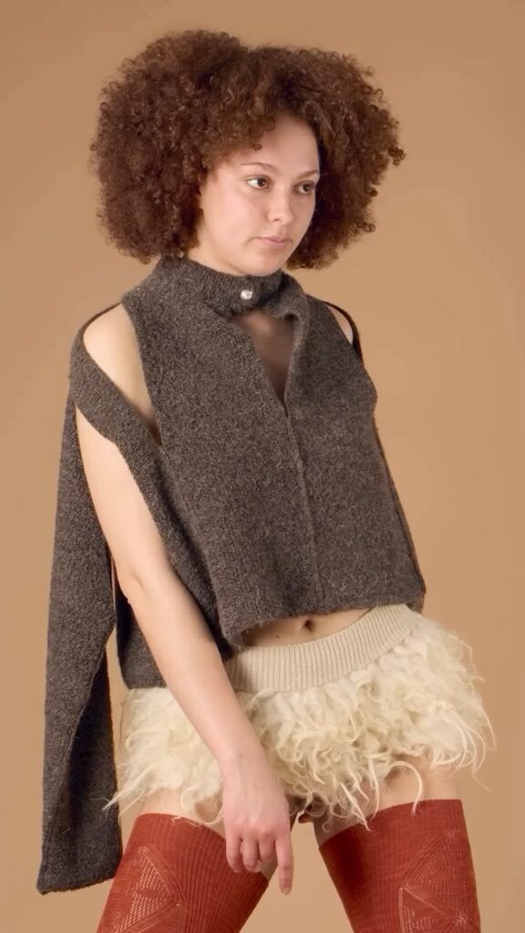 A person with curly hair wears a sleeveless gray wool top with open sides, a short cream-colored feathered skirt, and rust-colored thigh-high socks, standing against a beige background.