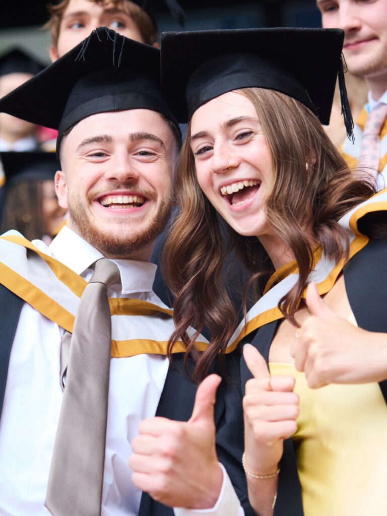 Two graduates in caps and gowns smile and give thumbs up while celebrating at a graduation ceremony. They appear joyful and excited, surrounded by other graduates in similar attire.