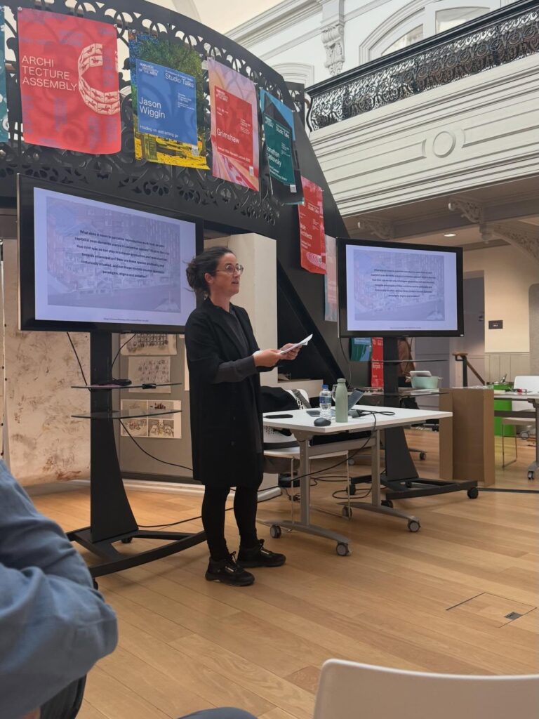 A woman in black stands and speaks in front of two large screens during a presentation at an architecture event, with colorful banners and architectural drawings displayed behind her.