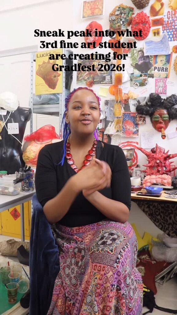 A smiling art student with blue braids and a colorful skirt sits in a vibrant art studio, surrounded by sculptures, masks, and various art materials. Text above reads, "Sneak peak into what 3rd fine art student are creating for Gradfest 2026!.