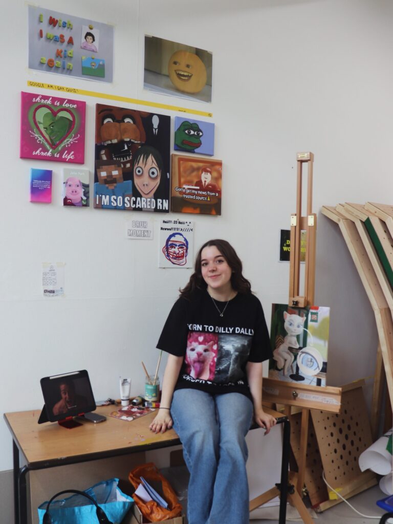 A young woman with long brown hair, wearing a black graphic t-shirt and flared jeans, sits on a desk in an art studio. Colorful meme-style artworks hang on the wall behind her, and an easel stands nearby.