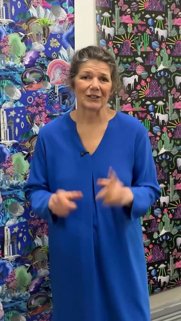 A woman in a bright blue dress stands in front of colorful, patterned wallpaper featuring animals and nature designs. She is speaking and gesturing with her hands.