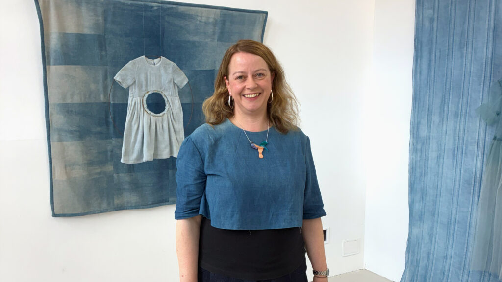 A woman with shoulder-length light brown hair, wearing a blue top and a necklace, smiles standing in front of blue textile art featuring a dress hung on a wall.
