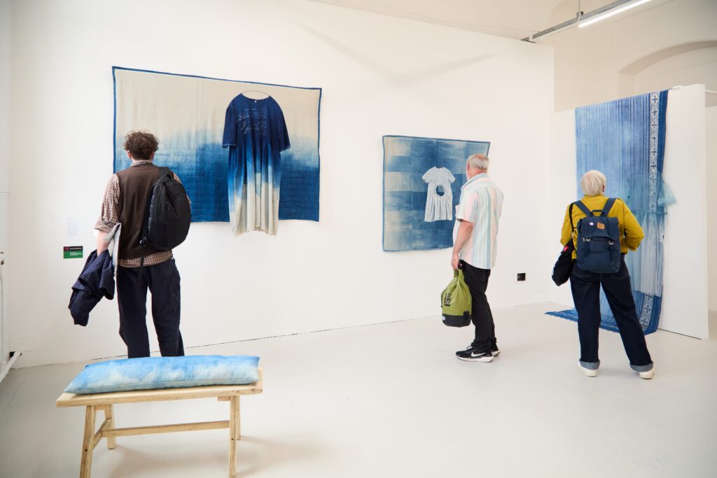 Three people view blue and white textile art displayed on a white gallery wall, including fabric panels and clothing, with a wooden bench in the foreground and another blue textile hanging to the right.
