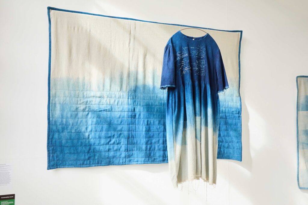 A blue and white quilt hangs on a wall with a blue and white dyed dress displayed in front, both featuring a gradient pattern from deep blue to white.