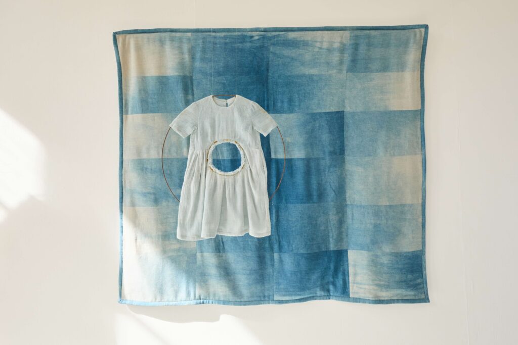 A small, white dress with a circular cutout at the waist hangs in front of a patchwork quilt made of blue and white fabric squares on a white wall.