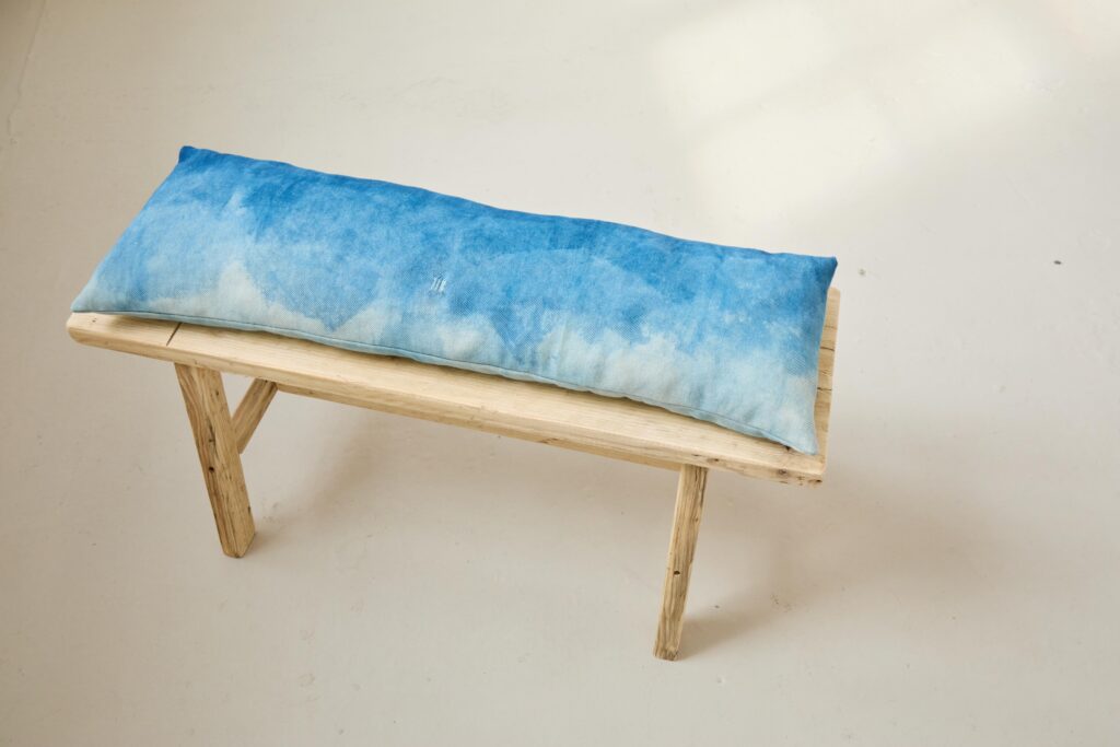 A long, rectangular bench cushion with a blue, watercolor-like pattern rests on a simple, light wooden bench set against a plain, off-white background.