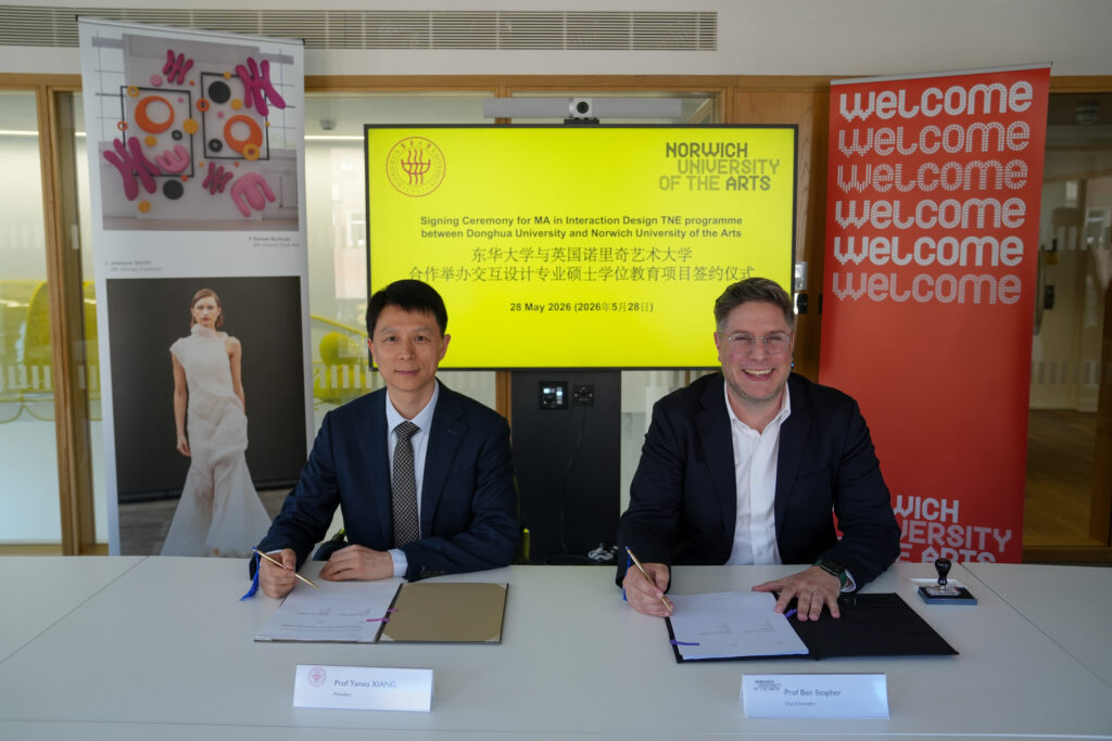 Two men in suits sit at a table, signing documents. Behind them, a screen displays “Norwich University of the Arts” and details of a partnership ceremony for the MA in Interaction Design. Welcome signs and a fashion poster are visible in the background.