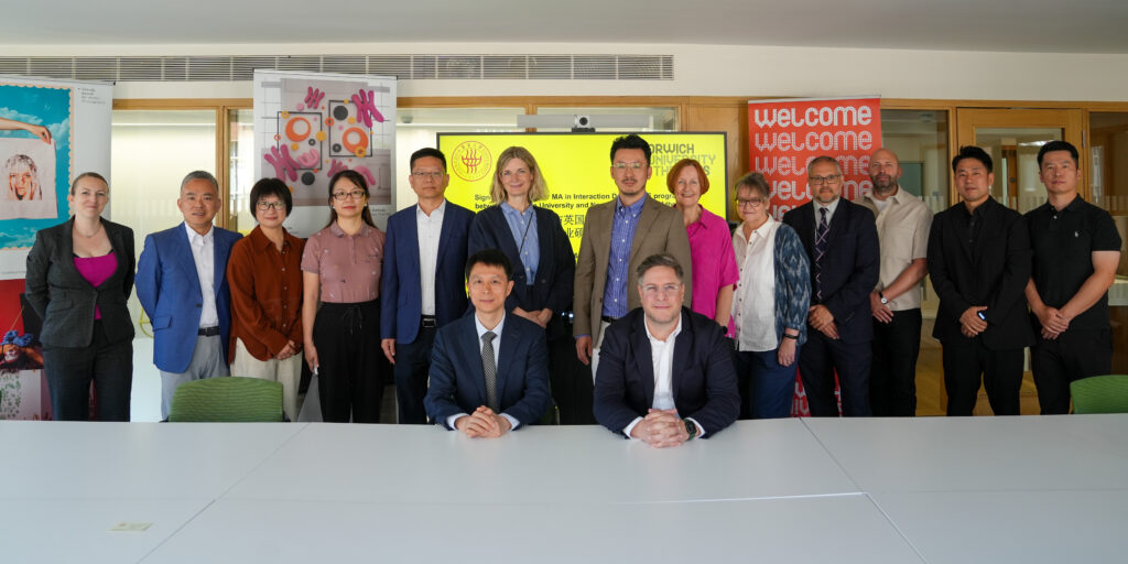 A group of fifteen professionally dressed people, including MA in Interaction Design graduates, pose in two rows—some standing and two seated at a conference table—in an office setting with welcoming posters and a presentation screen behind them.