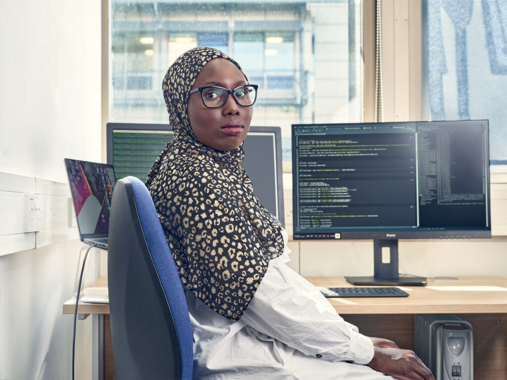 A woman wearing glasses and a patterned hijab sits at a desk with computer monitors displaying code in an office setting. She looks over her shoulder toward the camera.