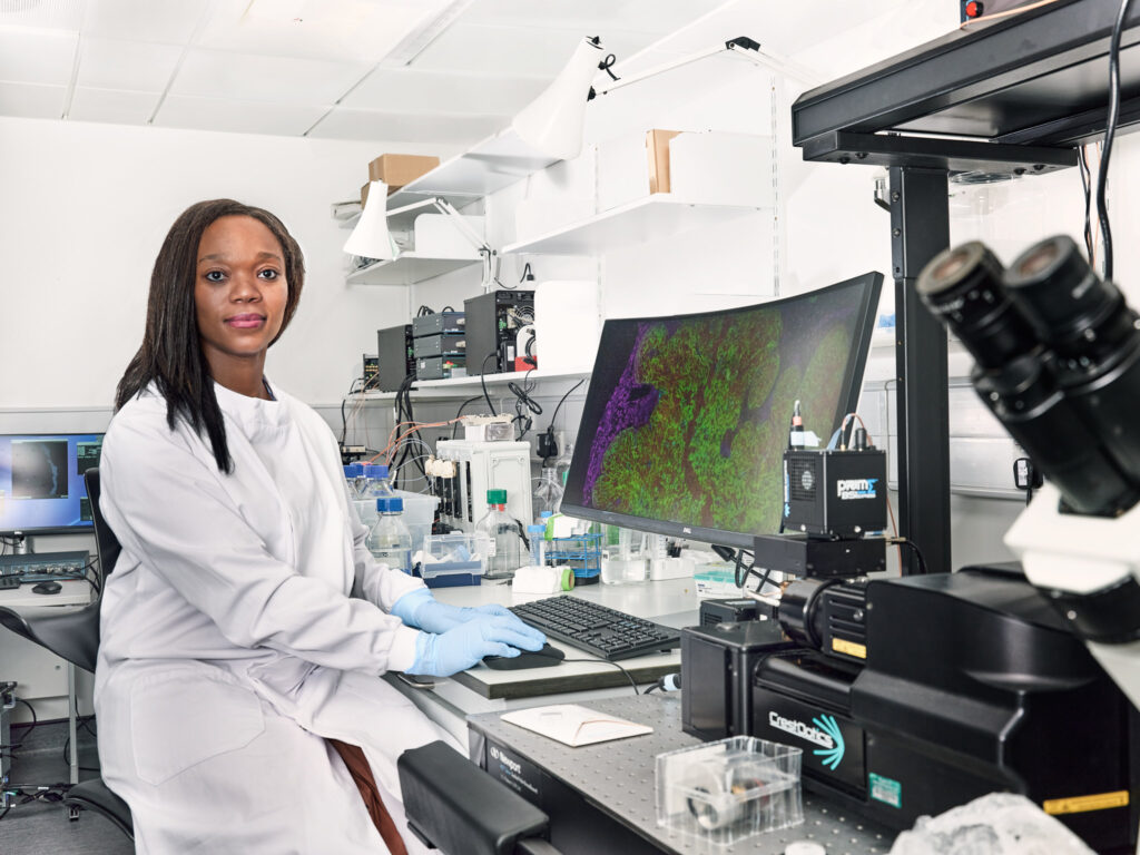 A scientist in a white lab coat and blue gloves sits at a desk in a laboratory, looking at the camera, with scientific equipment and a large computer screen displaying colorful cellular images.