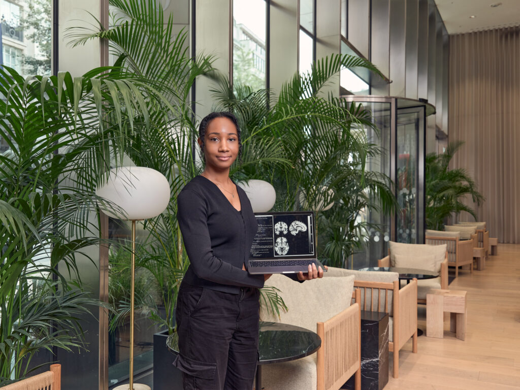 A person stands in a modern, plant-filled lounge holding a laptop displaying brain scan images. They wear a black outfit and look at the camera, with chairs and glass walls visible in the background.