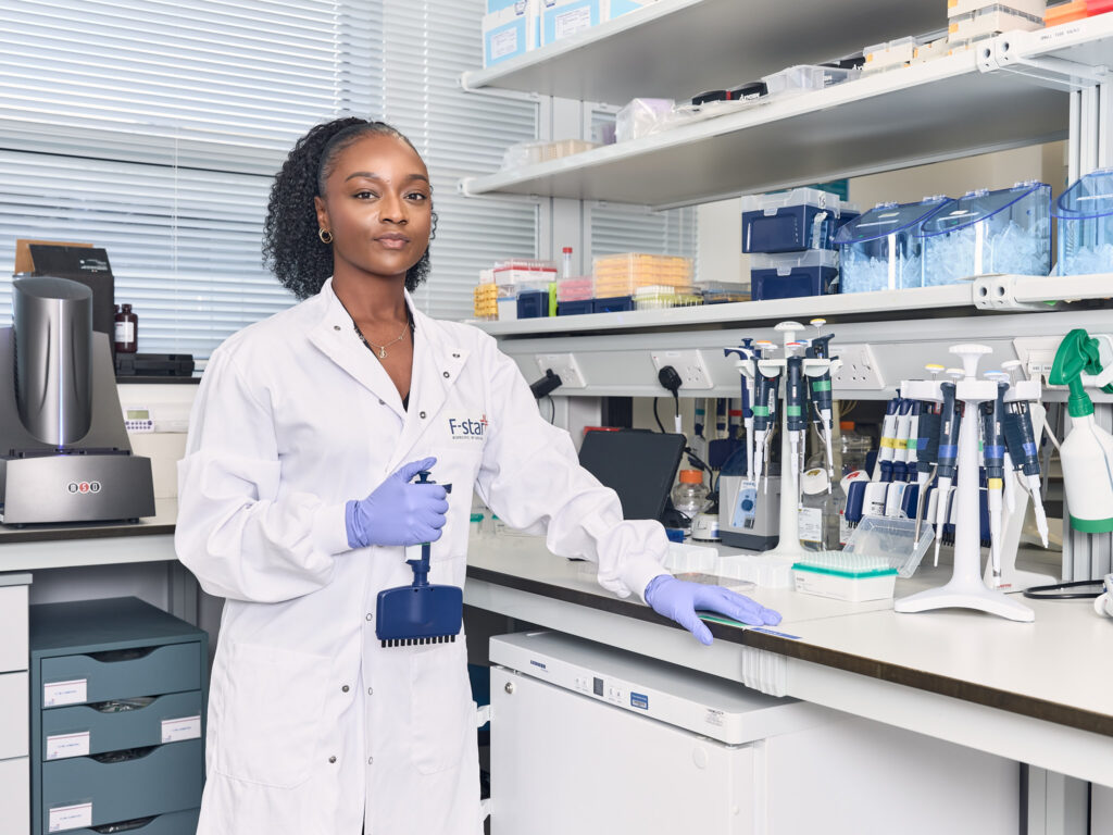 A scientist in a white lab coat stands in a laboratory, holding a pipette. She wears purple gloves and stands near a bench with lab equipment, tubes, and pipette racks, with shelves and scientific tools in the background.