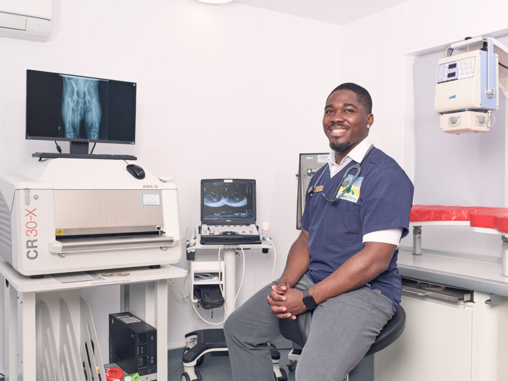 A smiling man in medical scrubs sits on a stool in a veterinary clinic, surrounded by medical equipment, including an X-ray of a dog's legs displayed on a monitor.