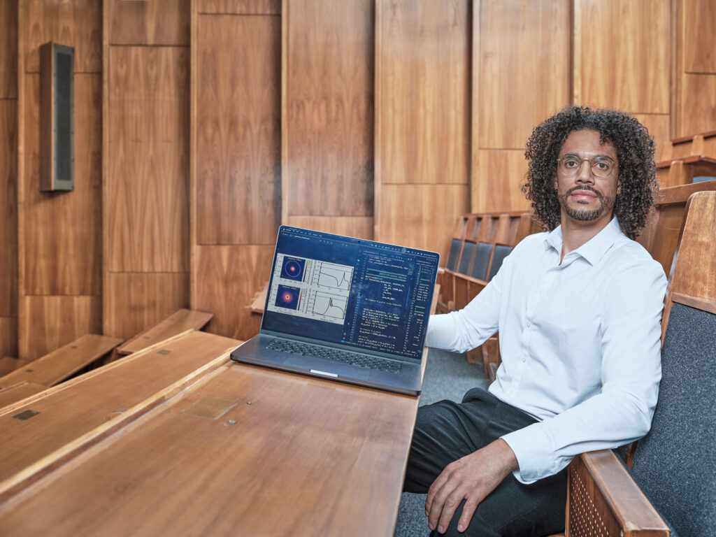 A person with curly hair and glasses sits in a wooden lecture hall, holding a laptop displaying data charts and code, looking directly at the camera.