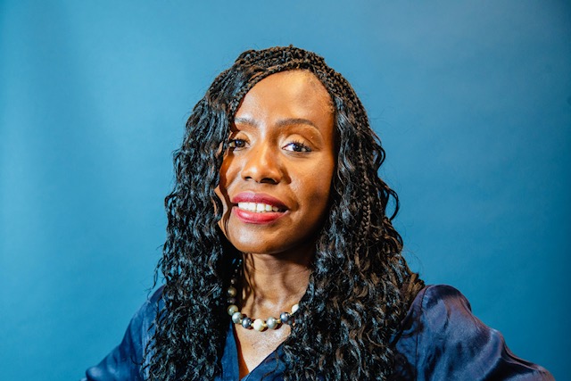 A woman with long curly hair smiles confidently against a solid blue background. She wears a dark blouse, a pearl necklace, and red lipstick.