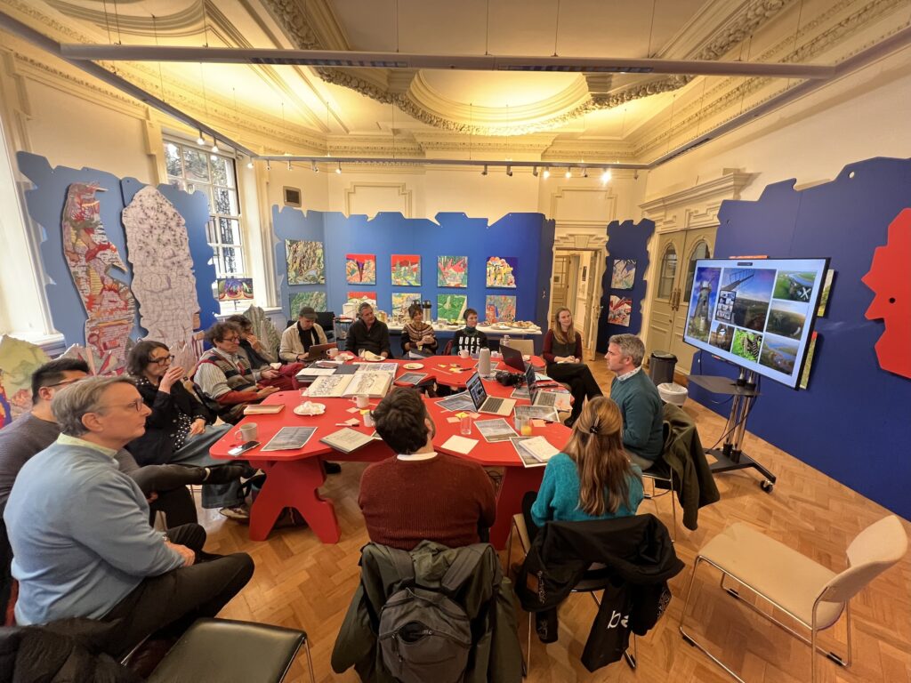 A group of people sit around a large red table in a bright, colorful meeting room with maps, artwork on blue walls, and a screen displaying images. Papers and laptops are spread across the table.