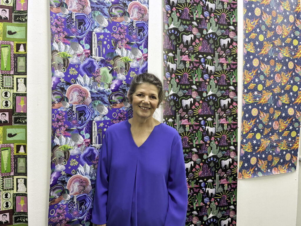 A woman in a blue blouse stands smiling in front of three panels of colourful, patterned fabric artworks displayed on a white wall.