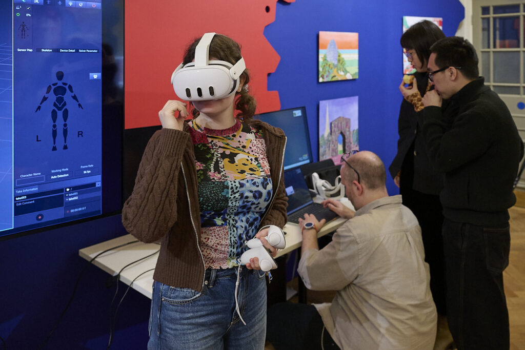 A woman wearing a VR headset stands in front of a screen displaying a digital interface. Three other people work at computers in the background, all in a room with colorful artwork on the walls.