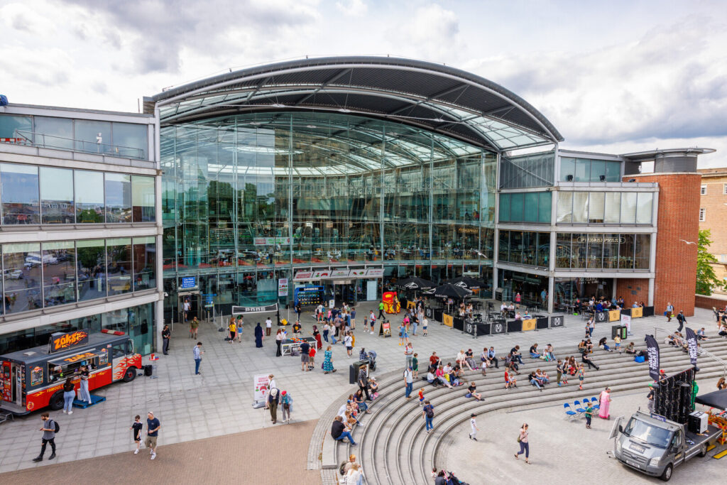 A modern glass-fronted building with a curved roof, busy with people walking and sitting on steps in a public plaza. Food trucks and market stalls are set up outside under a partly cloudy sky.