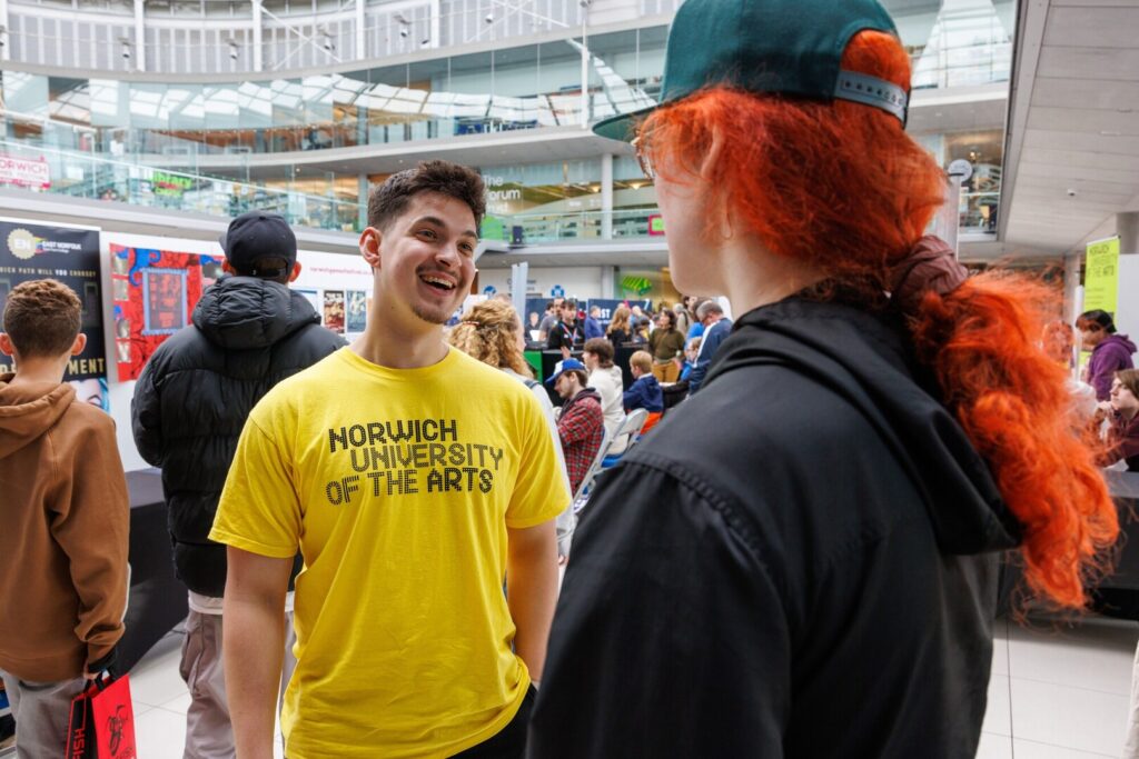 A smiling person in a yellow "Norwich University of the Arts" t-shirt talks to another person with long red hair, wearing a green cap, at a busy indoor event with people and displays in the background.
