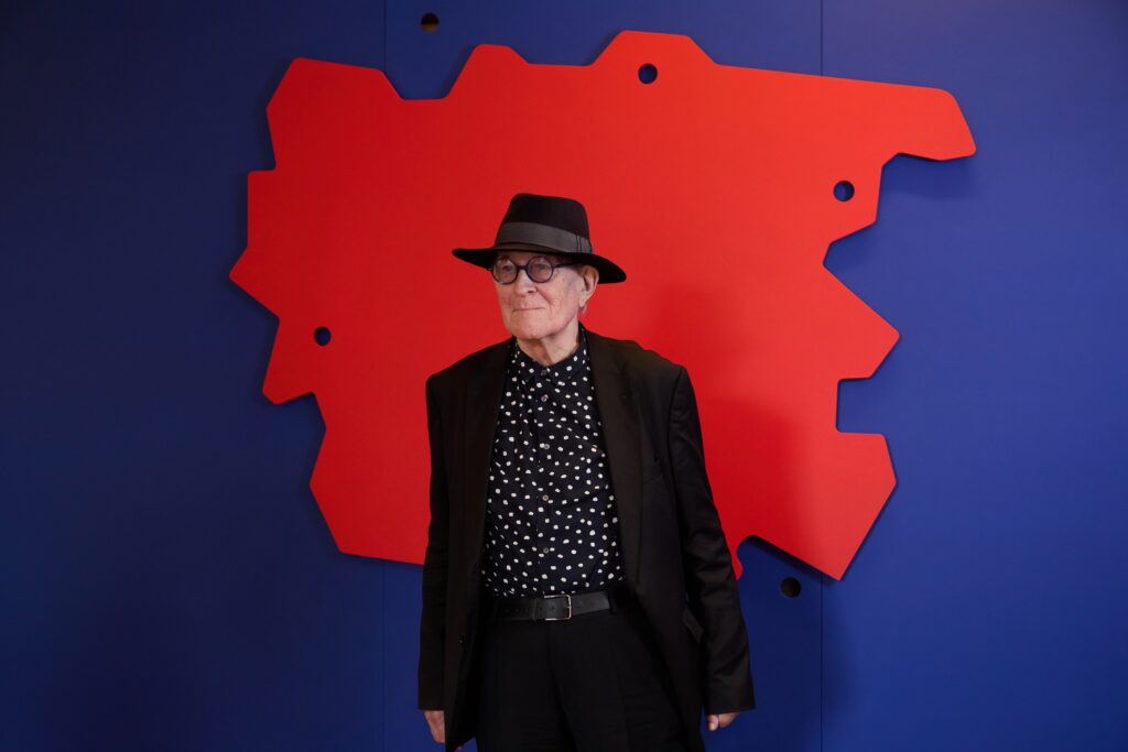 An older person wearing a black hat, polka dot shirt, and black jacket stands in front of a blue wall featuring an irregular red abstract shape with circular cutouts.