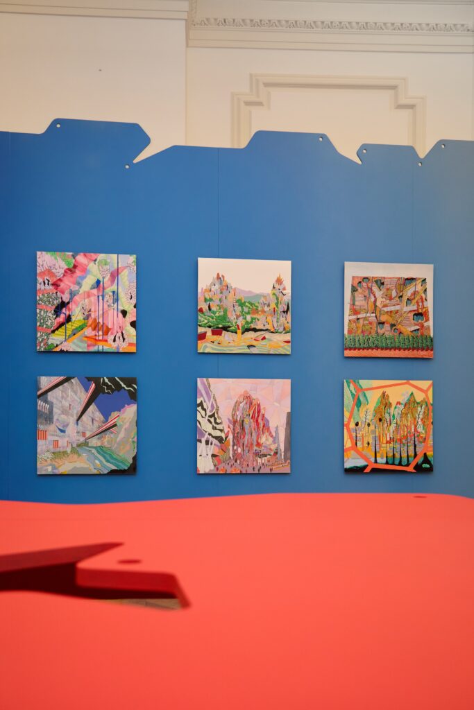 Six colorful abstract paintings are displayed on a blue wall in two rows of three. A pink and red surface is in the foreground, and decorative white molding is visible at the top of the cream-colored wall.