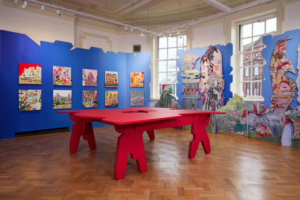 A brightly lit art gallery with parquet floors, blue walls displaying colorful modern paintings, large windows, and a bold, red sculptural table in the center of the room.