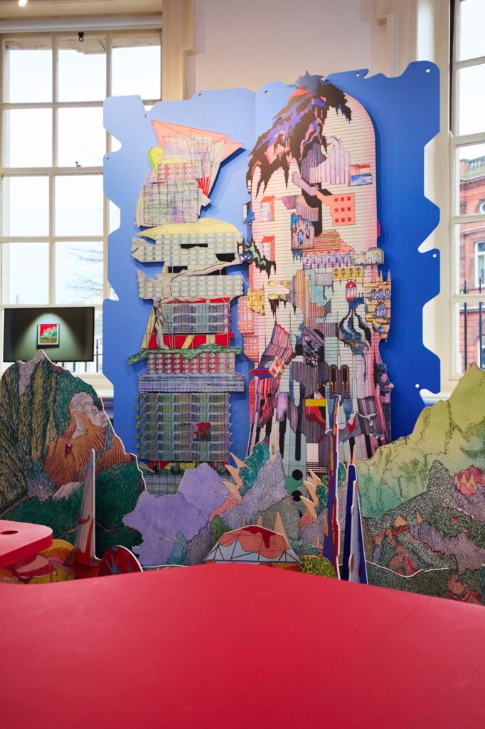 A colorful, abstract art installation features a large, intricate cityscape model with geometric shapes, buildings, and mountains, set against a blue backdrop in a bright gallery space with large windows.