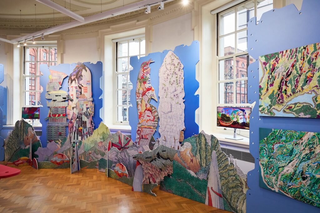 Colorful, cut-out collage artworks of fantastical cityscapes and mountains are displayed in a bright gallery with large windows and parquet floors; a monitor shows digital art on the right wall.