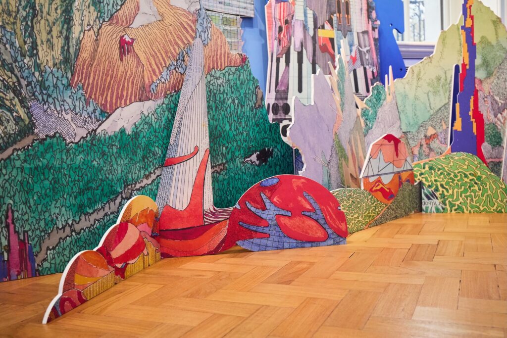 Colorful, cut-out cardboard pieces with abstract and landscape designs stand on a wood floor, forming a vibrant, layered art installation resembling mountains and hills with various textures and patterns.