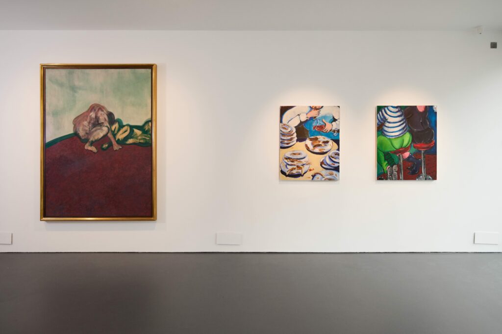 A white gallery wall displays three colorful paintings: a large framed painting on the left of a figure on a red surface, and two smaller paintings on the right showing food and people at a table. The floor is dark and the space is minimal.