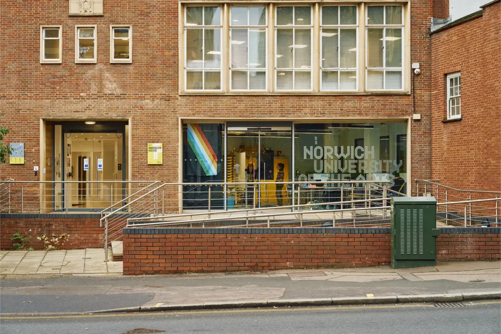 The entrance to a brick building with large windows, displaying "Norwich University" and a colorful diagonal stripe. A ramp leads to the door, and a green utility box is in front, next to the sidewalk and street.
