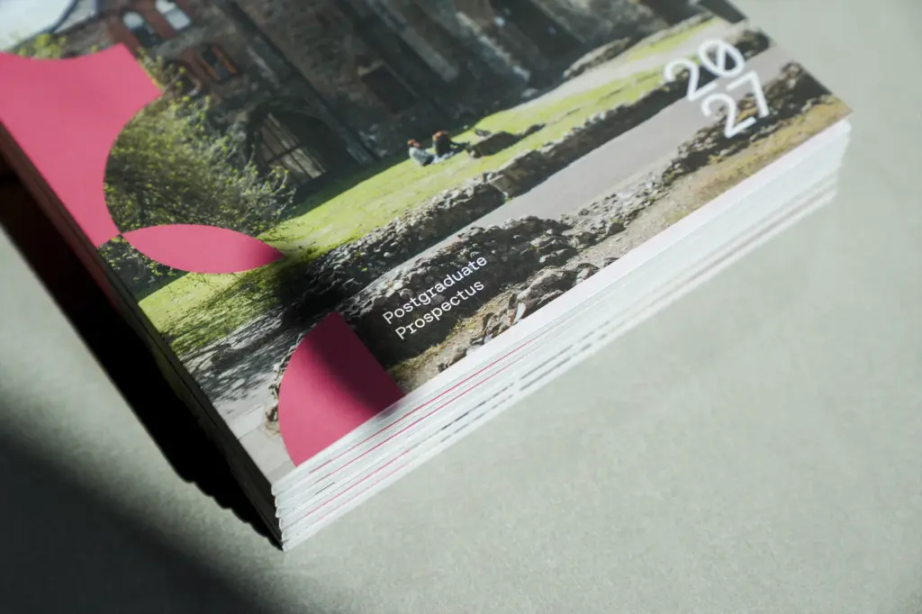 A close-up of a stack of brochures titled “Postgraduate Prospectus 2024/2025,” featuring a pink design and an outdoor campus scene with grass, stone walls, and a historic building.