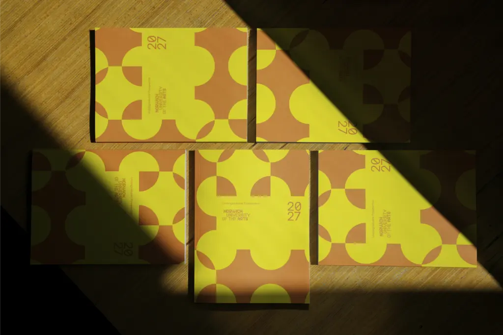 Five yellow and orange booklets with geometric patterns are arranged on a wooden surface, partially illuminated by a beam of sunlight, creating sharp contrasts of light and shadow across the covers.