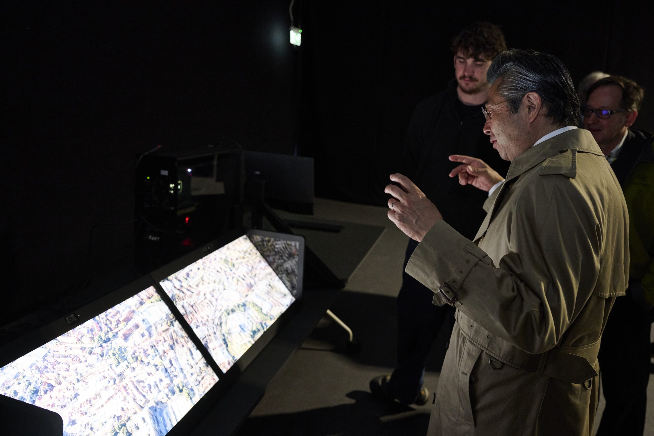 A man in a beige trench coat gestures toward two large screens displaying aerial images, while two other men stand nearby in a dark room.