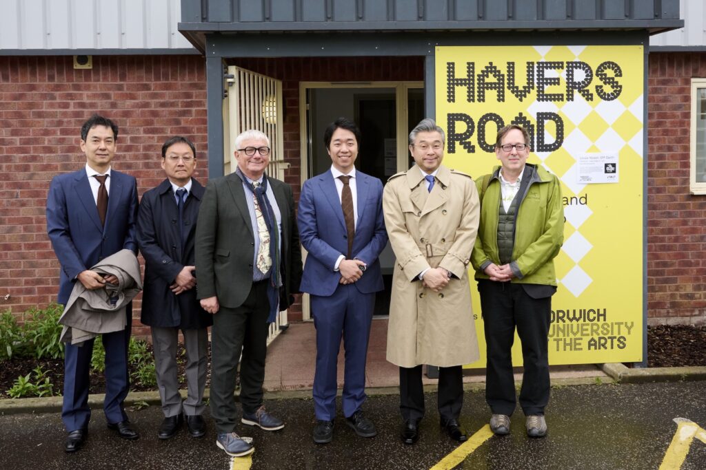 Six men in business attire stand smiling outside a building with a yellow "Havers Road" sign near the entrance, located at Norwich University of the Arts. The group appears posed for a photograph.