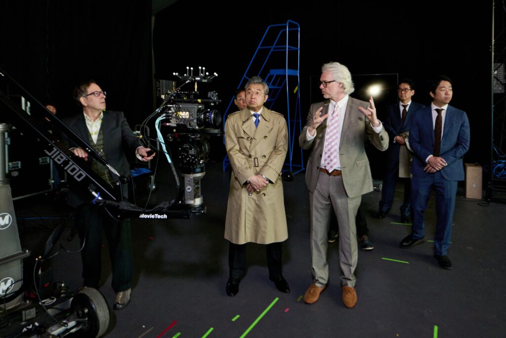 A group of men in suits and trench coats stand on a film set, surrounded by camera equipment. One man gestures while speaking, as others listen. Neon lines and tape markings are visible on the dark floor and walls.