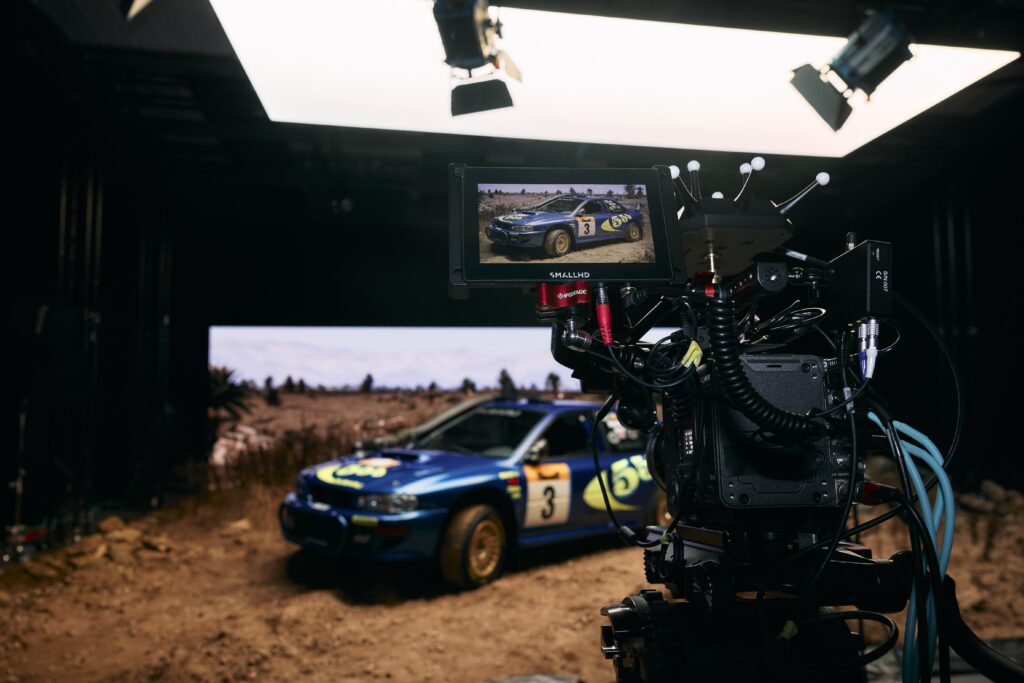 A camera films a blue rally car with racing decals in a studio setup, with the car visible on the camera’s monitor. Studio lights and a rocky ground create a realistic outdoor racing scene.