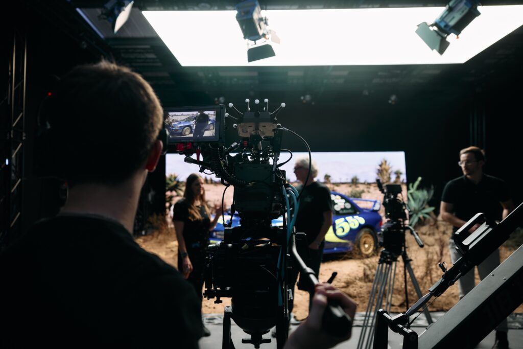 A camera operator films a group of people standing near a blue race car with the number 59 on its side, on a set with bright overhead lighting and filming equipment visible.