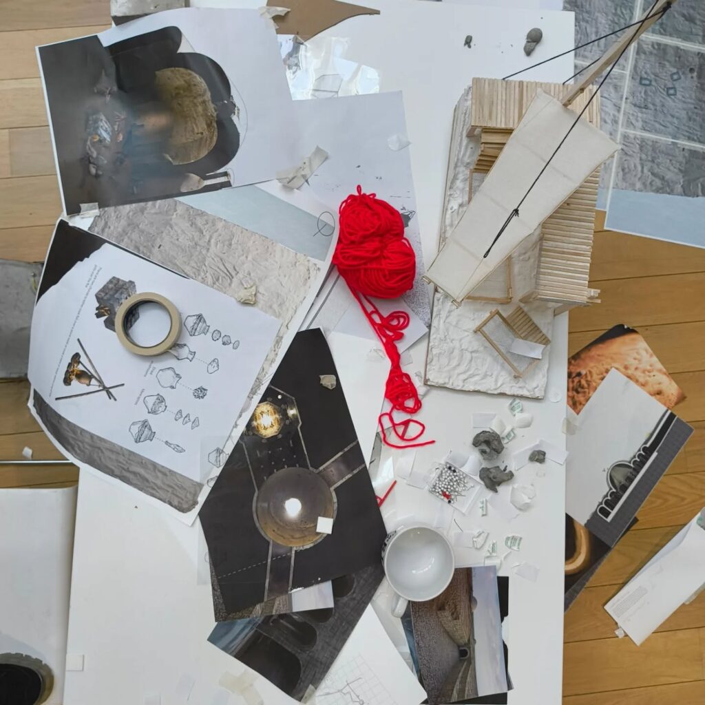 A cluttered workspace with architectural sketches, photographs, a red ball of yarn, a broken white cup, scattered paper, and a wooden architectural model on a white table over a wooden floor.