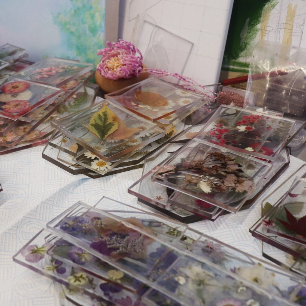 Stacks of clear resin coasters, each containing pressed flowers and leaves, are arranged on a patterned surface. In the background, art supplies and floral decorations are visible.