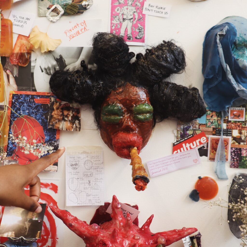 A hand points to a colorful collage on a wall, featuring a large sculpted mask with green eyes and a cigar-like object in its mouth, surrounded by various photos, drawings, notes, and textured art pieces.
